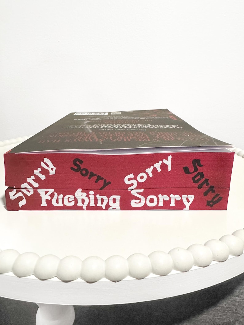 K&ouml;nnte beinhalten: Zwei B&uuml;cher auf einer wei&szlig;en Oberfl&auml;che gestapelt. Das obere Buch hat einen dunklen Einband. Das untere Buch hat einen roten Einband mit den Worten "Sorry" und "Fucking Sorry" in wei&szlig;er Schrift. Die B&uuml;cher stehen auf einem wei&szlig;en Perltablett.