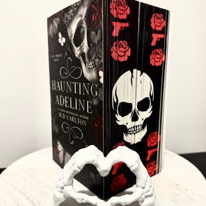 May include: A black book with a skull and roses design. The title of the book is "Haunting Adeline" by H.D. Carlton. The book is standing on a white surface.