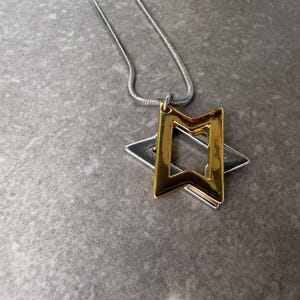 May include: A silver chain necklace with a pendant featuring a gold-colored rectangular shape over a silver star. The pendant is a geometric design with a modern aesthetic. The necklace is displayed on a gray surface.