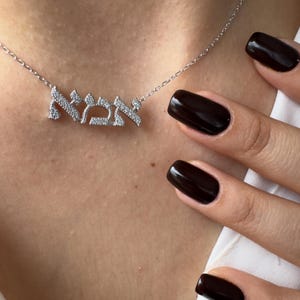 Könnte beinhalten: Eine silberne Halskette mit einem Anhänger mit hebräischen Schriftzeichen, verziert mit kleinen, klaren Steinen. Die Halskette hängt an einer feinen Kette. Der Hintergrund zeigt die Haut einer Person und dunklen Nagellack.