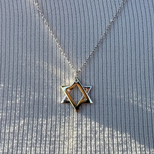 May include: A silver necklace with a Star of David pendant. The pendant has a two-tone design, with a gold-colored inner star and a silver-colored outer star. The necklace is displayed against a textured, light gray background.