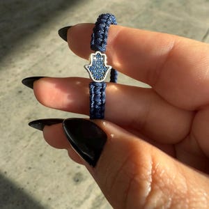 Puede incluir: Una pulsera trenzada azul con un dije de hamsa plateado. El dije está incrustado con cristales azules.