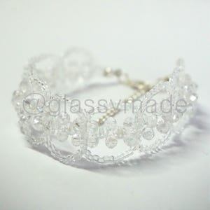May include: A clear beaded bracelet with a floral design. The bracelet is made of small, clear beads strung together to create a delicate, openwork pattern. The clasp is silver. The text "@glassymade" is visible.