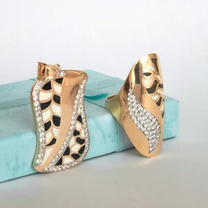 May include: Two gold-toned statement rings with a unique design. One ring features a pattern of black, white, and clear stones, while the other has a clear stone design. Both rings are displayed on a light blue box.