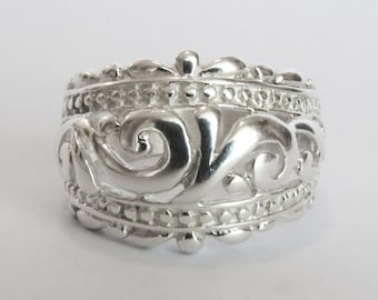 Puede incluir: Un anillo de plata con un diseño ornamental y ondulado. El anillo presenta un trabajo de volutas detallado y una superficie texturizada. La banda es ancha y el estilo es vintage o antiguo.