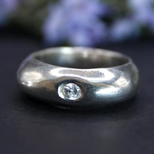 Könnte beinhalten: Ein silberner Ring mit einem abgerundeten Band und einem einzelnen, klaren Edelstein. Der Ring hat eine glatte, polierte Oberfläche und ein klassisches Design. Der Edelstein ist in einer Lünettenfassung gefasst.