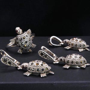 Könnte beinhalten: Ein silberfarbenes Schmuckset mit Schildkrötenmotiven. Das Set umfasst einen Ring und mehrere Anhänger, die alle mit kleinen, dunklen Steinen verziert sind. Der Panzer und der Körper der Schildkröte sind mit den Steinen detailliert, und der Ring hat einen roten Stein auf dem Kopf.