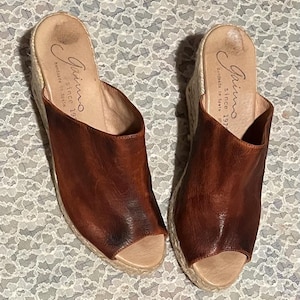 May include: A pair of brown leather espadrille wedges. The open-toe shoes have a woven jute sole and a leather upper. The brand name "Gaimo" and "handmade in Spain since 1978" are printed on the insole.