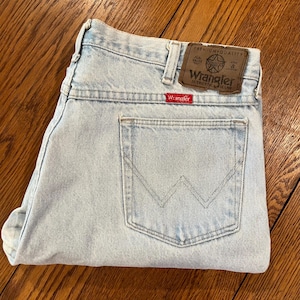 May include: Light blue Wrangler jeans with a classic five-pocket design. The jeans feature a brown leather patch with the Wrangler logo and a red Wrangler tag on the back pocket. The jeans are folded and appear unworn.