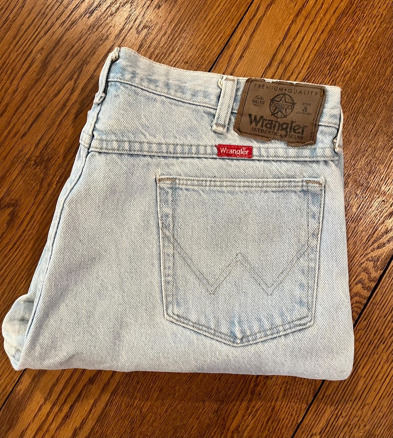 May include: Light blue Wrangler jeans with a classic five-pocket design. The jeans feature a brown leather patch with the Wrangler logo and a red Wrangler tag on the back pocket. The jeans are folded and appear unworn.