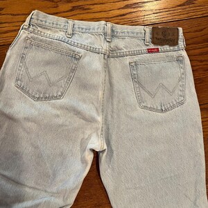 May include: Light blue denim jeans with the Wrangler logo on the back pocket and a brown leather patch. The jeans have a classic five-pocket design and a straight leg fit. The back pockets feature the iconic Wrangler "W" stitching.