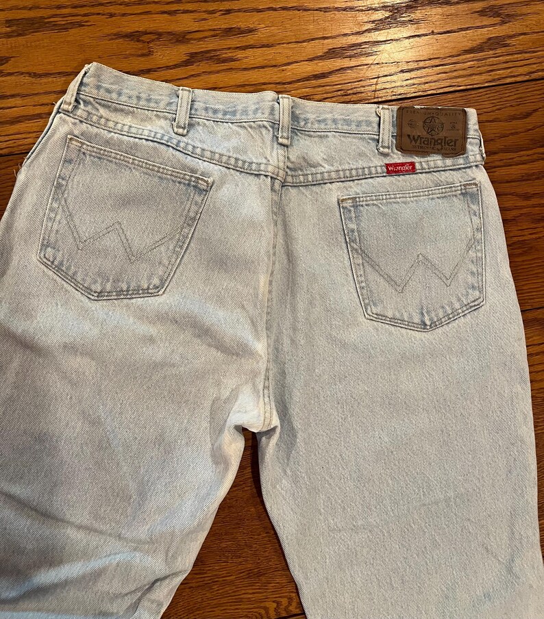 May include: Light blue denim jeans with the Wrangler logo on the back pocket and a brown leather patch. The jeans have a classic five-pocket design and a straight leg fit. The back pockets feature the iconic Wrangler "W" stitching.