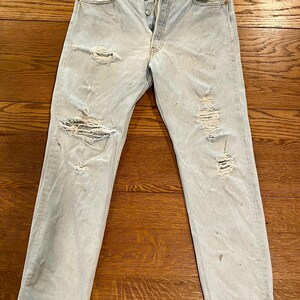 May include: Light blue denim jeans with distressed details, including rips and fading. The jeans have a button fly and classic five-pocket styling. The garment is laid flat on a wooden surface.