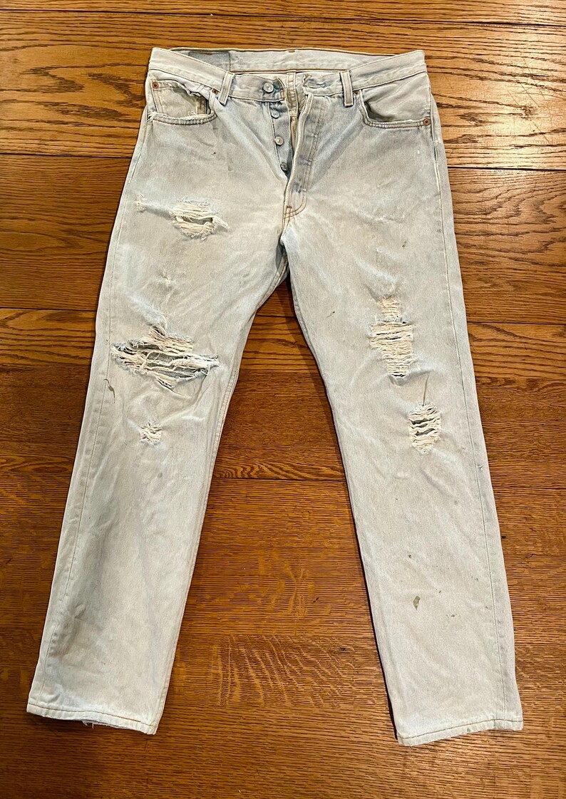 May include: Light blue denim jeans with distressed details, including rips and fading. The jeans have a button fly and classic five-pocket styling. The garment is laid flat on a wooden surface.