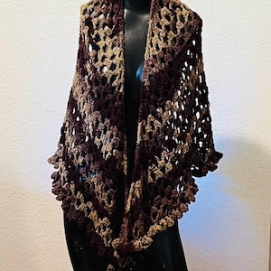 May include: A handmade crochet shawl in shades of brown and tan, displayed on a black mannequin. The open weave design features a scalloped edge, creating a textured and stylish accessory. This wrap is perfect for adding warmth and style.