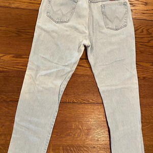 May include: Light blue denim jeans with the Wrangler logo on the back pocket. The jeans have a classic five-pocket design and a straight leg cut. The jeans are laid flat on a wooden surface.