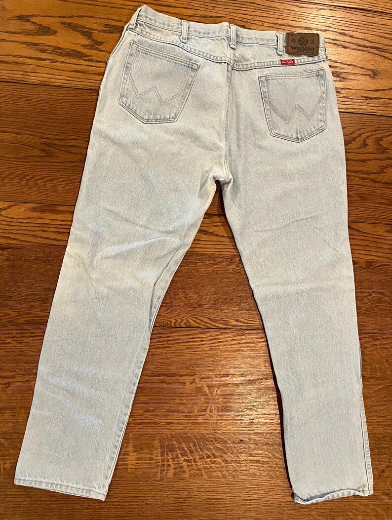 May include: Light blue denim jeans with the Wrangler logo on the back pocket. The jeans have a classic five-pocket design and a straight leg cut. The jeans are laid flat on a wooden surface.