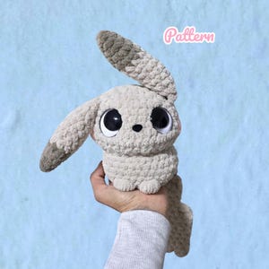 May include: A plush crochet toy shaped like a rabbit with large black eyes, a small black nose, and long ears. The toy is made of soft, beige yarn with darker brown tips on the ears. The word Pattern is written in pink script in the upper right corner.