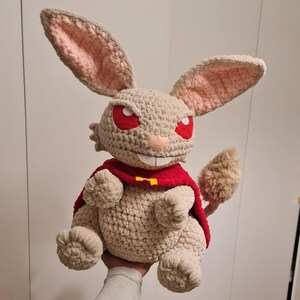 Pu&ograve; includere: Bambola coniglio all'uncinetto fatta a mano con mantello rosso e occhi rossi. Il coniglio ha grandi orecchie con interno rosa, corpo beige e coda soffice. Il mantello ha un emblema giallo.