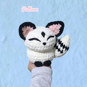 May include: A white plush toy with a crochet texture, featuring large ears with pink inner details, black accents on the ears and feet, and a bushy tail with a black and white checkered pattern. The word Pattern is written in pink above the toy.