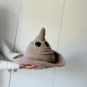 Könnte beinhalten: Ein handgefertigter, taupefarbener Häkelhut mit großen, ausdrucksstarken Augen. Der Hut hat eine breite Krempe und eine spitze Oberseite, die einem skurrilen Charakter ähnelt. Dieses einzigartige Accessoire ist perfekt für Kostüme oder Cosplay.