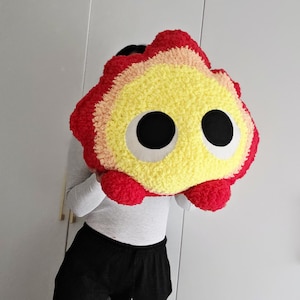 May include: A large, plush, cartoon-like creature with a vibrant color scheme. The creature features a red, yellow, and peach-colored exterior with large, black, circular eyes. The plush toy is designed with a soft, textured fabric.