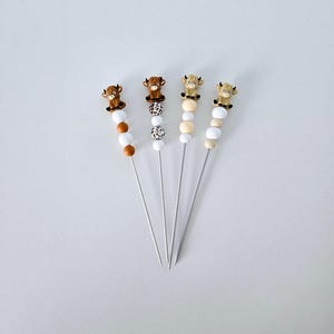 May include: Four decorative skewers with a cow figurine on top. The skewers have a series of beads in white, brown, and patterned designs. The cow figurines are brown and tan. The skewers are arranged on a white surface.