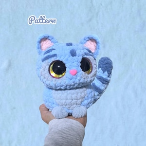 Könnte beinhalten: Ein hellblaues und graues gehäkeltes Katzen-Plüschtier mit großen, runden, dunklen Augen mit goldfarbenen Iris, einer kleinen rosa Nase und Streifenmustern auf Kopf und Schwanz. Das Wort Pattern ist in weißem Text in der oberen linken Ecke gedruckt.