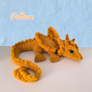 May include: A mustard yellow crocheted dragon plush toy with a long, curved tail, textured wings, and dark, glossy eyes. The word Pattern is printed in a light orange font in the upper left corner of the image above the dragon figure.