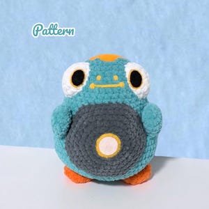 May include: A teal crochet plush toy with a dark gray circular belly patch featuring a white center. The creature has large white eyes with black pupils, an orange patch on its head, and orange feet. The word Pattern is printed in white.