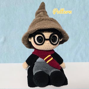 May include: A crochet doll featuring a tan face, black hair, and large black glasses. The doll wears a brown pointed hat, a black robe, and a maroon scarf with yellow stripes. The word Pattern is written in yellow text in the upper right corner.