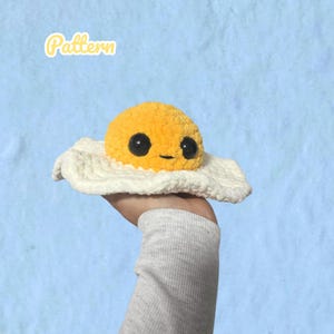 Puede incluir: Un juguete de peluche tejido a ganchillo con forma de huevo frito, con una yema amarilla brillante y una base blanca. La yema tiene dos grandes ojos de seguridad de plástico negro y una pequeña boca negra bordada. La palabra Pattern está en amarillo.