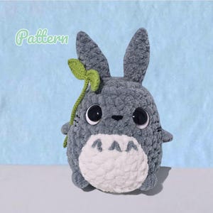 May include: A gray crocheted plush toy with large black eyes, a white belly featuring three gray chevron markings, and a green leaf attached to the side of its head. The word Pattern is written in light green script in the upper left corner.