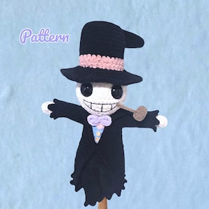 May include: A crocheted doll featuring a white head with large black eyes, a wide stitched grin, and a brown pipe. The doll wears a black top hat with a pink band, a black coat with a light blue vest, two small buttons, and a light purple bow tie.