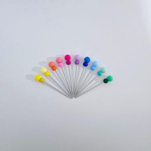 Könnte beinhalten: Eine Fächeranordnung von Nähstiften mit silbernen Schäften und bunten, runden Köpfen. Die Köpfe sind in einem Regenbogen von Farben gehalten, darunter Gelb, Orange, Rosa, Lila, Blau und Grün. Auf weißem Hintergrund.
