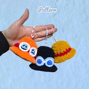May include: Three miniature crochet bucket hats hanging from beaded loops. One is orange with white and blue goggles, one is black with blue goggles, and one is yellow with a red band. The word Pattern is printed in white at the top of the image.