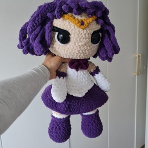以下が含まれることがあります： セーラームーンをテーマにした手作りのぬいぐるみ。人形は紫色のウィッグ、金のティアラ、白と紫の衣装が特徴です。人形は人の手で持たれています。