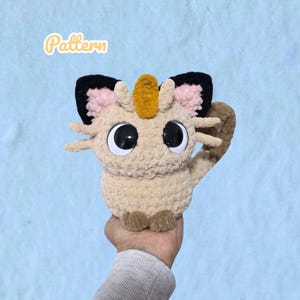 Peut inclure: Une peluche crochetée beige avec de grands yeux noirs et blancs, des oreilles bordées de noir avec des centres roses, une breloque ovale dorée sur le front et des moustaches. Le mot Pattern est écrit en lettres cursives blanches au-dessus.