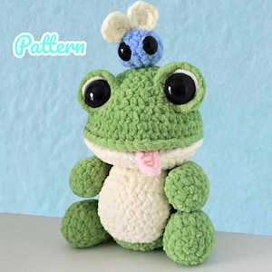 Könnte beinhalten: Ein gehäkeltes grünes Frosch-Stofftier mit großen schwarzen Augen, einem cremefarbenen Bauch und einer kleinen herausgestreckten rosa Zunge. Eine kleine blau-cremefarbene Biene sitzt auf dem Kopf des Frosches. Das Wort Pattern steht in Weiß.