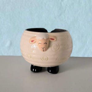 Könnte beinhalten: Ein cremefarbener Keramik-Blumentopf in Schaf-Form mit schwarzem Innenraum und schwarzen Füßen. Das Schaf hat ein lächelndes Gesicht mit rosa Ohren und einem weißen, wolligen Kopf. Der Körper weist geprägte Wirbelmuster auf.