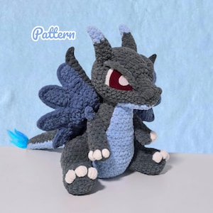 May include: A dark gray and light blue crocheted dragon plush toy with large dark blue wings, white claws, and red eyes. The word Pattern is written in white script in the upper left corner. The creature has a long tail with a bright blue tip.