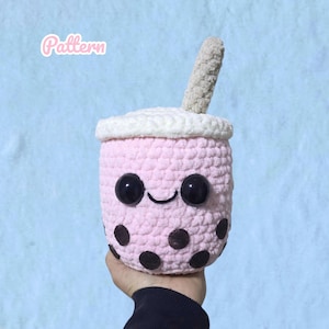 Könnte beinhalten: Ein gehäkeltes Bubble-Tea-Plüschtier in Hellrosa mit weißem Deckel und beigem Strohhalm. Das Spielzeug hat zwei große schwarze Sicherheitsaugen aus Kunststoff und ein aufgesticktes schwarzes Lächeln. Dunkelbraune Filzkreise sind außen angebracht.