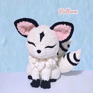 May include: A white crocheted plush toy featuring black accents on the ears, paws, and tail, with pink inner ears and a black diamond-shaped marking on the forehead. The plush has closed eyes and the word Pattern is written in the upper right corner.