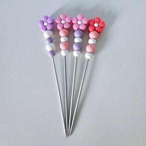 Peut inclure: Quatre piques à cocktail décoratives avec des fleurs violettes, roses et rouges. Chaque pique a une série de perles blanches, violettes et roses. Les piques sont en métal et sont disposées sur une surface blanche.
