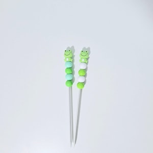 Op de afbeelding: Twee decoratieve spiesen met een kikkerontwerp. Elke spies heeft een reeks groene en witte kralen, bekroond met een lachende groene kikker. De spiesen zijn gemaakt van metaal en zijn ideaal voor hapjes of cocktails.