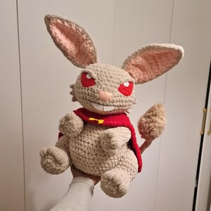 Pu&ograve; includere: Una bambola coniglio all'uncinetto fatta a mano con grandi orecchie, occhi rossi e un mantello rosso. Il peluche &egrave; beige con accenti rosa all'interno delle orecchie e una coda soffice. Il coniglio ha un sorriso malizioso e tiene in mano un mantello rosso con una chiusura dorata.