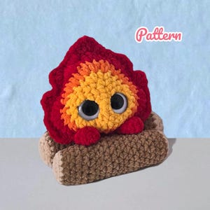Könnte beinhalten: Ein gehäkeltes Plüschtier in Flammenform mit großen schwarz-weißen Kunststoffaugen aus rotem und orangefarbenem Garn, das auf zwei braunen gehäkelten Holzscheiten ruht. Das Wort Pattern steht in weißer Schrift in der oberen rechten Ecke.