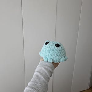 以下が含まれることがあります： 黒い目をした水色の手編みのタコぬいぐるみ。手作りの玩具は白い背景に掲げられています。タコは丸い頭と8つの小さな触手を持っています。ぬいぐるみは柔らかく、質感のある糸で作られています。
