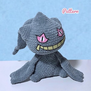 May include: A grey crocheted plush toy featuring a pointed head, long drooping ears, and wide pink eyes with black pupils. The character has a wide, yellow, toothy grin. The word Pattern is printed in pink script in the upper right corner.