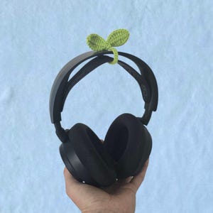 Op de afbeelding: Zwarte over-ear koptelefoon met een groen gehaakt blad detail. De koptelefoon wordt tegen een lichtblauwe achtergrond gehouden. Het blad is lichtgroen.